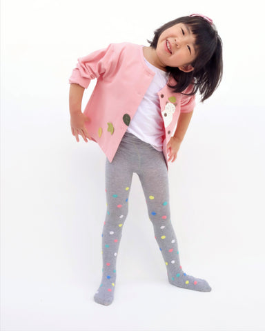 A playful girl standing, smiling, and wearing a pink relaxed-fit cotton cat jacket with an appliqué peekaboo cat and falling leaves in the front. There are buttons on the jacket. 