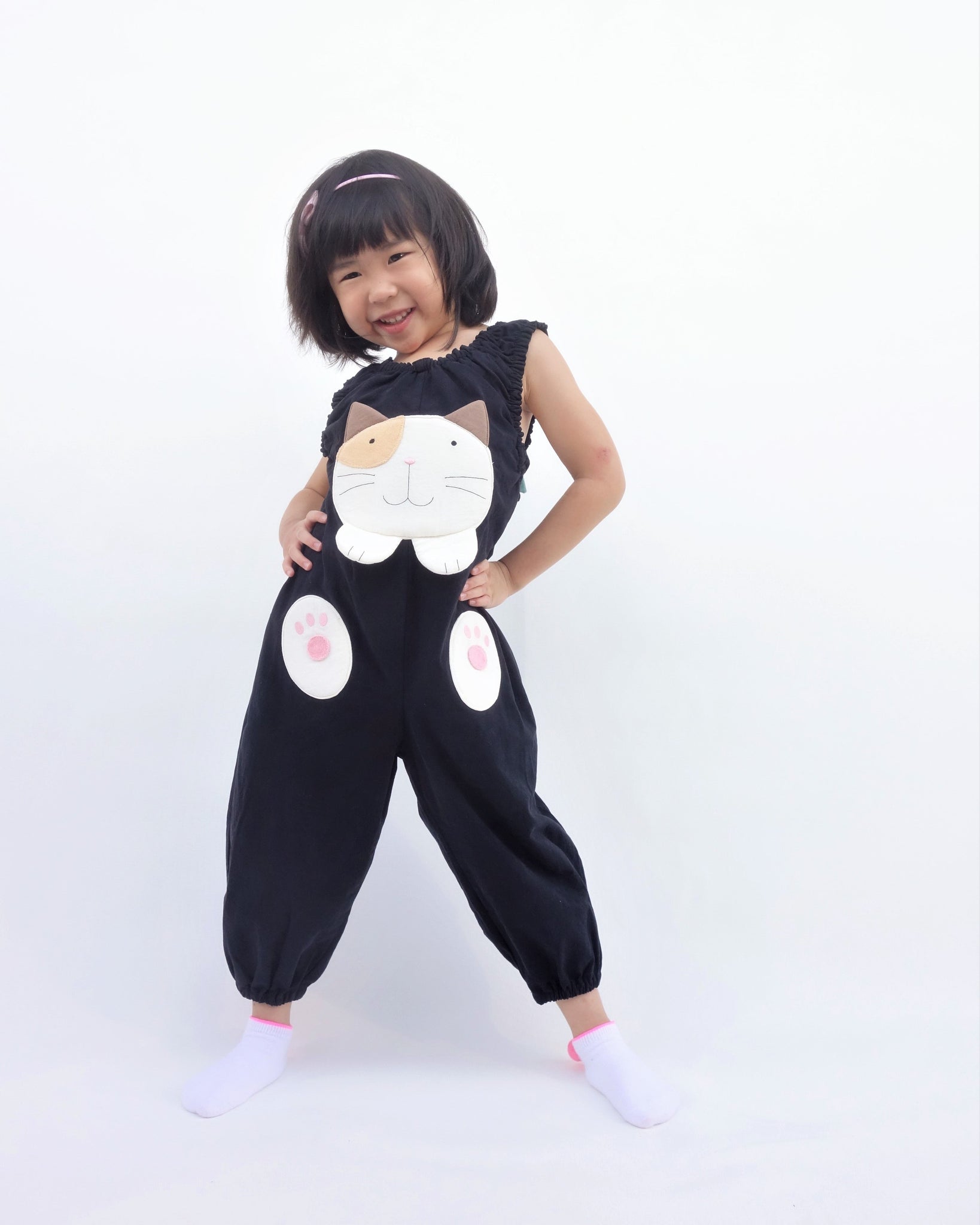 Girl wearing black, cat-themed jumper with large, soft cat face pocket and cat paws in the front, two little fish friends swim on the back, sewn appliqué, embroidery details and side pockets in front view.