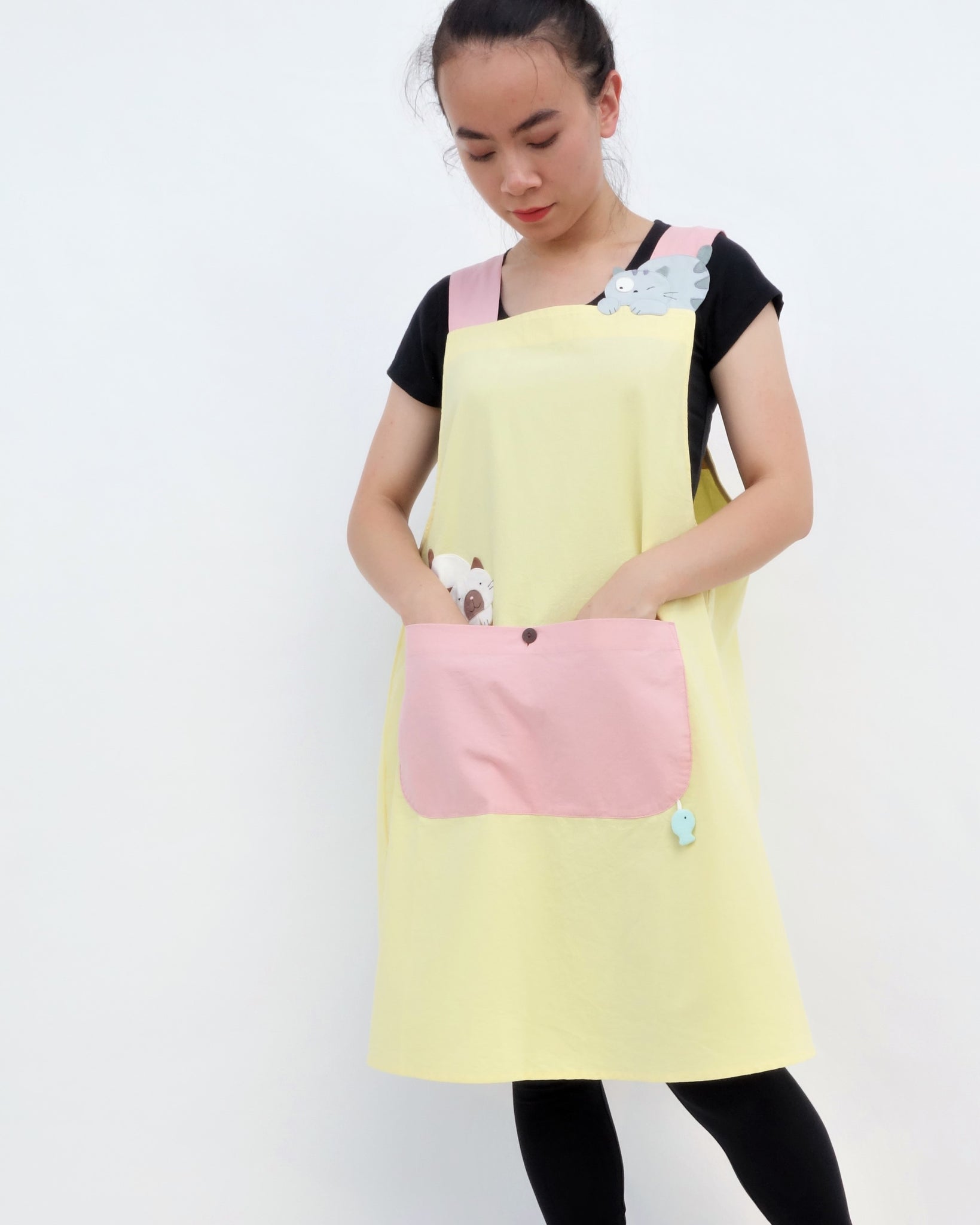 Woman wearing pink and yellow color-blocked apron with cat appliqué, large front pocket, in front close-up view.