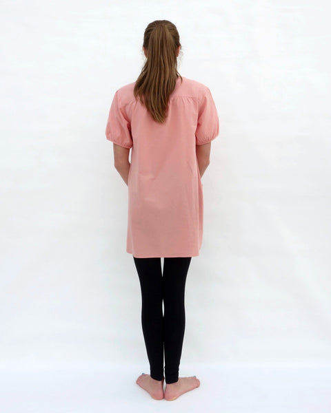 A woman standing, wearing a pink cotton cat-themed tunic dress for women with black leggings underneath, back facing. The tunic dress has puffed sleeves and slits on the side. 