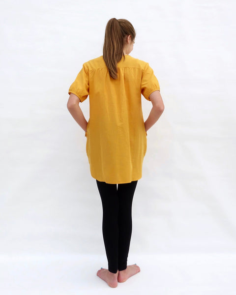 A woman standing, wearing a yellow cotton cat-themed tunic dress for women with black leggings underneath, back facing. The tunic dress has puffed sleeves and slits on the side. 