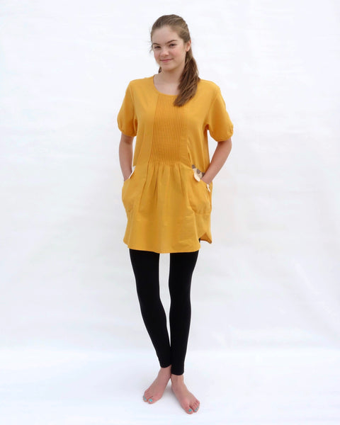 A woman with hands in the pockets, wearing a yellow cotton cat-themed tunic dress for women with two cats/kittens on the pockets and black leggings underneath. The tunic dress has puffed sleeves and slits on the side. 