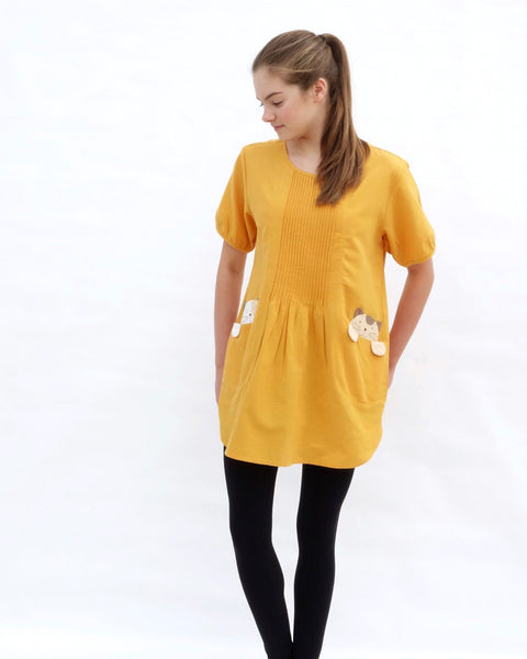 A woman with hands on the sides, wearing a yellow cotton cat-themed tunic dress for women with two cats/kittens on the pockets and black leggings underneath. The tunic dress has puffed sleeves and slits on the side. 