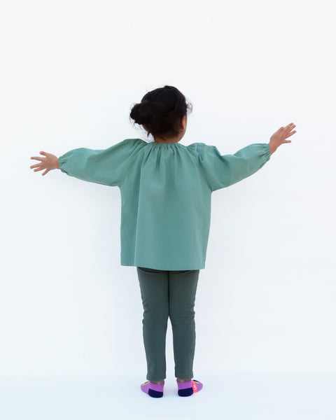 A girl wearing a loose fitting cat blouse in sage green that is light and airy with elastic around the neck, puffed sleeves, elastic stretch near the wrists in back view.