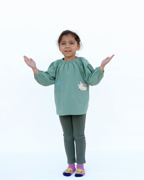 A girl gesturing and wearing a loose fitting cat blouse in sage green that is light and airy with elastic around the neck, sewn-on cat appliqué, embroidery details, puffed sleeves, elastic stretch near the wrists, and two front pockets in front view