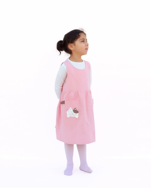Cute girl wearing pink sleeveless cat dress with kittens and kitten tails on the pockets in side view.
