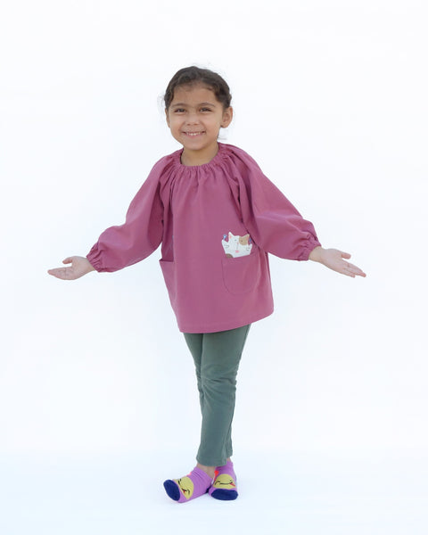 A cross-legged girl wearing a loose fitting cat blouse in dull rose that is light and airy with elastic around the neck, sewn-on cat appliqué, embroidery details, puffed sleeves, elastic stretch near the wrists, and two front pockets in front view.