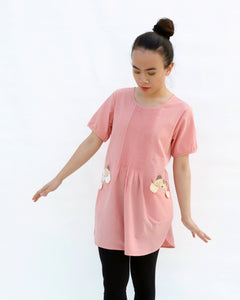 A woman standing, wearing a pink cotton cat-themed tunic dress for women with two cats/kittens on the pockets and black leggings underneath. The tunic dress has puffed sleeves and slits on the side. 