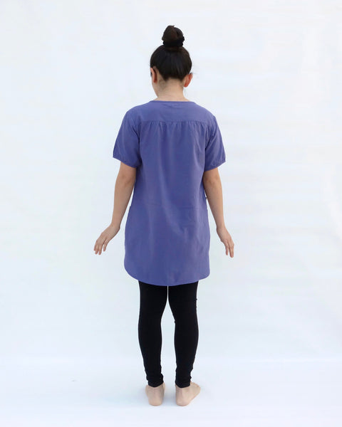 A woman standing, wearing a purple cotton cat-themed tunic dress for women with black leggings underneath, back facing. The tunic dress has puffed sleeves and slits on the side. 