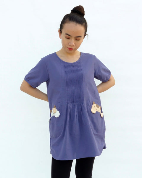 A woman with hands on hips, wearing a purple cotton cat-themed tunic dress for women with two cats/kittens on the pockets and black leggings underneath. The tunic dress has puffed sleeves and slits on the side. 