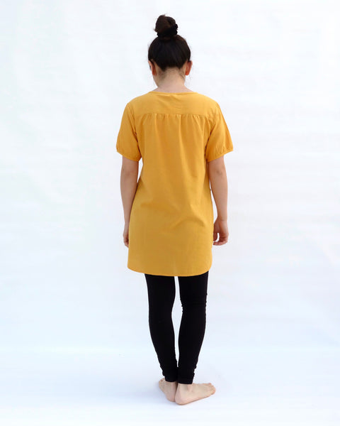 A woman standing, wearing a yellow cotton cat-themed tunic dress for women with black leggings underneath, back facing. The tunic dress has puffed sleeves and slits on the side. 