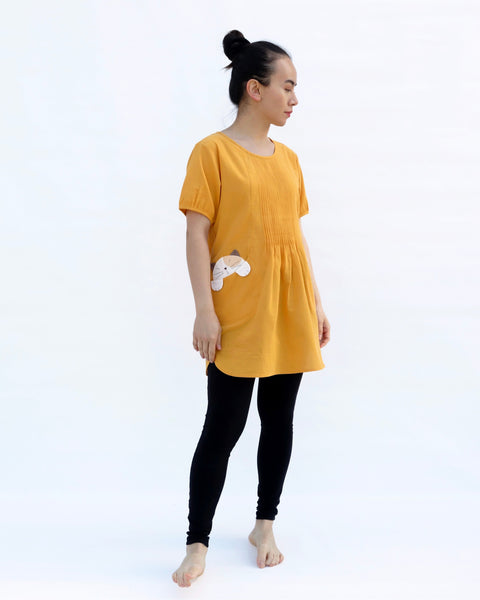 A woman with hands at her sides, wearing a yellow cotton cat-themed tunic dress for women with two cats/kittens on the pockets and black leggings underneath, side view. The tunic dress has puffed sleeves and slits on the side. 