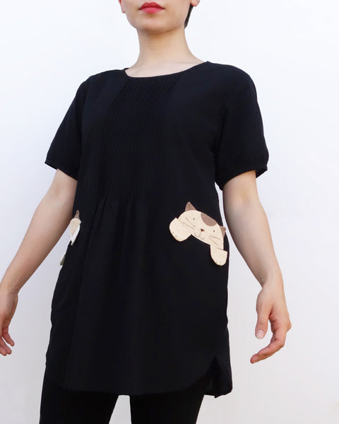 Woman wearing a black cotton cat-themed tunic dress for women with two cats/kittens on the pockets. The tunic dress has puffed sleeves and slits on the side. 