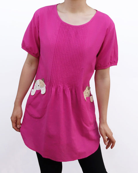 Woman wearing a hot pink/fuchsia cotton cat-themed tunic dress for women with two cats/kittens on the pockets. The tunic dress has puffed sleeves and slits on the side. 