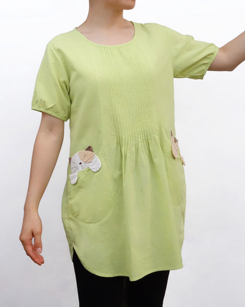 Woman wearing a light green cotton cat-themed tunic dress for women with two cats/kittens on the pockets and black tights leggings underneath. The tunic dress has puffed sleeves and slits on the side. 