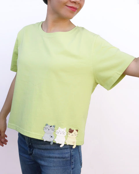 Women wearing a Cat Crop Top in green with bottom hem at the waist, three-cat appliqué on the front, one cat appliqué on the back, embroidery details, crew neck, and short sleeves in front close-up view. 