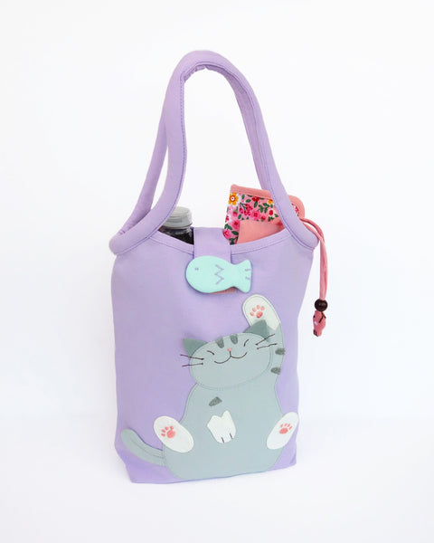 Cat Bottle-Tote in lilac with cotton interior, nylon lining, cat appliqué, embroidery detail, large main compartment, Velcro flap closure, sturdy double padded handles, squared off bottom for holding books, wallet, water bottle, pencil pouch, or umbrella.