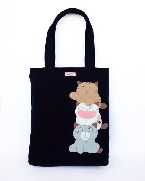 A  black cat-themed canvas tote bag with three appliqué cats playfully stacked on top of each other
